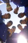 Низкий угол обзора многонациональной команды бейсболистов-мужчин, готовящихся к игре, мотивирующих друг друга в тесноте, смотрящих вниз на камеру, в солнечный день — стоковое фото