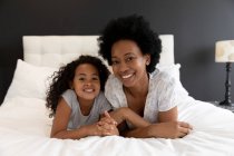 Retrato de uma mulher afro-americana e sua jovem filha relaxando no quarto juntos, deitados lado a lado na cama e sorrindo para a câmera — Fotografia de Stock