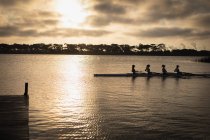 Vista lateral de un equipo de remo de cuatro mujeres caucásicas entrenando en el río, remando en una concha de carreras al amanecer, con la luz del sol reflejada en las ondas del agua y un embarcadero en primer plano - foto de stock