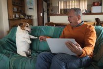 Передній погляд на старшого кавказького чоловіка відпочиває вдома у своїй вітальні, сидить на дивані розмовляючи зі своїм собакою і за допомогою ноутбука. — стокове фото