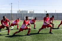 Equipe multiethnique de cinq hommes un joueur de football de côté portant un entraînement de bande d'équipe sur un terrain de sport au soleil, échauffement étirement. — Photo de stock