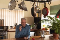 Вид спереди пожилого кавказца, расслабляющегося дома, сидящего за прилавком на кухне со смартфоном и улыбающегося — стоковое фото