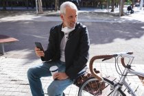 Старший кавказский мужчина днем бродит по улицам города в маске против коронавируса, ковид 19, сидит на скамейке запасных, держит чашку кофе на вынос и пользуется смартфоном. — стоковое фото