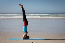 Vista laterale di una donna attraente caucasica, indossa vestiti sportivi, pratica yoga, in piedi nella sua testa in posizione yoga, sulla spiaggia soleggiata. — Foto stock
