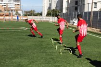 Multi-ethnische Mannschaft von männlichen Fünfer-Fußballern, die auf einem Sportplatz in der Sonne ein Strip-Training absolvieren und sich beim Laufen beim Springen über Hürden aufwärmen. — Stockfoto