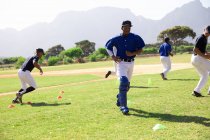 Вид сбоку на многонациональную группу бейсболистов-мужчин, тренирующихся со своим тренером на игровом поле, тренирующихся, бегущих, в солнечный день — стоковое фото