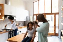 Передній вид на молоду афроамериканку вдома на кухні, сидячи на кухонному острові, а її батько прив'язує волосся, мати стоїть на задньому плані приготування їжі. — стокове фото