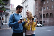 Передній погляд на щасливу кавказьку пару і на вулиці міста протягом дня, тримаючи чашечки кави, використовуючи смартфон і посміхаючись. — стокове фото