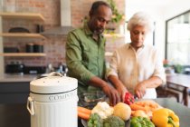 Feliz pareja jubilada afroamericana jubilada en casa, preparando comida, cortando verduras y sonriendo en su cocina, en casa juntos aislándose durante la pandemia del coronavirus covid19 - foto de stock