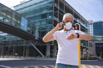 Старший кавказский мужчина днем бродит по улицам города в маске против коронавируса, ковид 19, используя смартфон и смартчасы. — стоковое фото