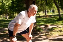 Vista lateral de un hombre mayor caucásico maduro haciendo ejercicio en un parque en un día soleado, recostado sobre sus rodillas, tomando un descanso - foto de stock