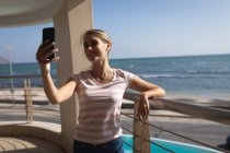 Kaukasische Frau steht auf einem Balkon, hält ein Smartphone in der Hand und macht ein Selfie. Soziale Distanzierung und Selbstisolierung in Quarantäne. — Stockfoto