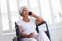 Una anciana afroamericana jubilada en casa, sentada en una silla de ruedas con pijama frente a una ventana en un día soleado hablando en un teléfono inteligente y sonriendo, auto aislándose durante la pandemia del coronavirus covid19 - foto de stock