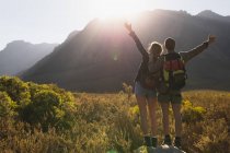 Vista posteriore da vicino di una coppia caucasica che si diverte durante una gita in montagna, camminando su un campo sotto le montagne, in piedi su una roccia insieme, alzando le mani, in una giornata di sole — Foto stock