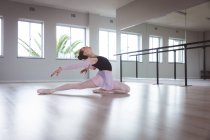 Atractiva bailarina de ballet caucásica con el pelo rojo estirándose hacia atrás, preparándose para una clase de ballet en un estudio brillante, centrándose en su ejercicio, sentada en el suelo. - foto de stock