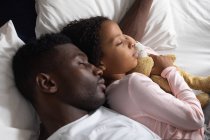 Niña afroamericana y su padre se distancian socialmente en casa durante el encierro de cuarentena, pasan tiempo juntos, abrazan mientras duermen. - foto de stock