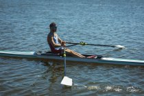 Vista lateral de un remo macho caucásico entrenando y remando en el río, sosteniendo remos remos y sentados en un bote de remos en un día soleado - foto de stock