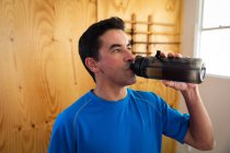 Vista frontale ravvicinata di un allenatore di judo maschio di razza mista concentrato che beve acqua da una bottiglia di plastica, in piedi in palestra facendo una pausa in un allenamento. — Foto stock