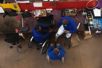 Grupo de trabajadores afroamericanos discapacitados en un taller en una fábrica que fabrica sillas de ruedas, sentados en un banco de trabajo ensamblando partes de un producto, dos sentados en sillas de ruedas, uno usando muletas - foto de stock