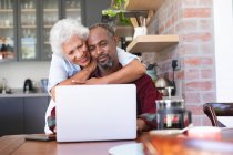 Una feliz pareja afroamericana jubilada en una mesa en su comedor, usando una computadora portátil juntos, el hombre sentado y la mujer de pie detrás y abrazándolo, ambos sonriendo - foto de stock