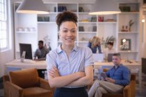 Porträt einer fröhlichen Geschäftsfrau mit gemischter Rasse, die in einem modernen Büro arbeitet, in die Kamera blickt und lächelt, während ihre Kollegen im Hintergrund arbeiten — Stockfoto