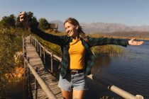 Передня частина жінки з Кавказу добре проводить час у поїздці в гори, стоячи на мосту, тримаючи смартфон і роблячи селфі в сонячний день. — стокове фото