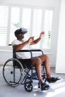 Ein älterer afroamerikanischer Rentner sitzt an einem sonnigen Tag in Unterwäsche im Rollstuhl vor einem Fenster und benutzt ein VR-Headset mit ausgestreckten Armen vor sich, um sich während der Coronavirus-Pandemie selbst zu isolieren 19 — Stockfoto