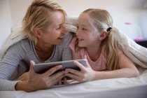 Front view of a Caucasian woman enjoying family time with her daughter at home together, lying in bed in their bedroom, smiling and looking at each other, using tablet computer — Stock Photo