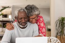 Una pareja afroamericana mayor pasa tiempo en casa juntos, distanciamiento social y aislamiento en cuarentena durante la epidemia de coronavirus covid 19, sentado en una mesa, usando un ordenador portátil, abrazando y sonriendo - foto de stock
