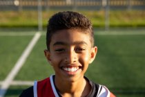 Портрет крупним планом щасливого змішаної раси хлопчика-футболіста в командній смузі, що стоїть на ігровому полі на сонці, дивлячись на камеру і посміхаючись — стокове фото