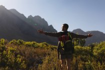 Rückansicht eines kaukasischen Mannes, der sich bei einem Ausflug in die Berge amüsiert, auf einem Feld unter den Bergen steht und die Aussicht genießt, die Arme weit ausgebreitet, an einem sonnigen Tag — Stockfoto