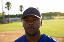 Retrato de um jogador misto de beisebol masculino, vestindo um uniforme de equipe e um boné, em pé em um campo de beisebol, olhando para a câmera — Fotografia de Stock