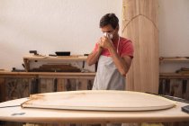 Homem caucasiano fabricante de pranchas de surf trabalhando em seu estúdio, vestindo um avental protetor, colocando uma máscara facial se preparando para polir uma prancha de surf . — Fotografia de Stock