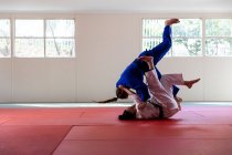 Seitenansicht von zwei jugendlichen kaukasischen und gemischten Judokas, die blau-weiße Judogis tragen und während eines Sparrings in einer Sporthalle Judo üben. — Stockfoto