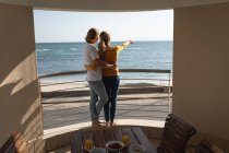 Kaukasisches Paar, das auf einem Balkon steht, sich umarmt und aufs Meer zeigt. Soziale Distanzierung und Selbstisolierung in Quarantäne. — Stockfoto
