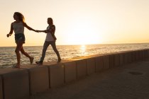 Кавказская пара, идущая по набережной у моря на закате, держась за руки, женщина ведет. Романтическая пляжная пара — стоковое фото