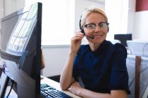 Porträt einer kaukasischen Geschäftsfrau mit kurzen blonden Haaren, die in einem modernen Büro arbeitet, am Schreibtisch sitzt, Headset trägt und in die Kamera blickt — Stockfoto