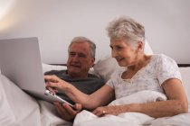 Primer plano de una feliz pareja de ancianos caucásicos jubilados en casa sentados en la cama en su dormitorio, hablando y usando una computadora portátil juntos y sonriendo, pareja aislando durante coronavirus covid19 pandemia - foto de stock