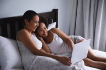 Vista laterale di una coppia mista di donne che si rilassano a casa in camera da letto, si siedono sul letto usando un computer portatile insieme e sorridono — Foto stock