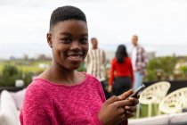 Portrait d'une Afro-Américaine traînant sur une terrasse sur le toit par une journée ensoleillée, regardant la caméra, souriant, tenant un smartphone, avec des gens parlant en arrière-plan — Photo de stock