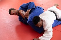 Vue de face à angle élevé d'un entraîneur de judo masculin de race mixte et d'un judoka masculin de race mixte adolescent portant du judoka bleu et blanc, pratiquant le judo lors d'un entraînement dans un gymnase. — Photo de stock