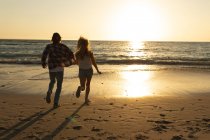 Кавказька пара біжить на пляжі під час заходу сонця, тримаючись за руки і дивлячись на море. — стокове фото