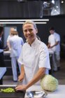 Retrato de uma cozinheira caucasiana cortando legumes, olhando para a câmera e sorrindo, com outros chefs cozinhando ao fundo. Aula de culinária em uma cozinha de restaurante. — Fotografia de Stock