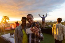 Vista frontale di un uomo di razza mista e una donna afroamericana appesi su una terrazza sul tetto con un cielo al tramonto, prendendo un selfie, con persone che tengono una bandiera americana sullo sfondo — Foto stock