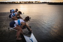 Vue latérale en angle élevé d'une équipe d'aviron de quatre femmes caucasiennes debout sur une jetée et plaçant un bateau dans la rivière en préparation de l'entraînement à l'aviron — Photo de stock