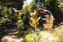 Kaukasische Frau mit Schürze und ihre Tochter genießen die gemeinsame Zeit in einem sonnigen Garten, betrachten gemeinsam Pflanzen und tragen eine Auswahl an Pflanzen in Körben. — Stockfoto