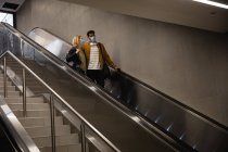 Vista frontale ad angolo basso di una coppia caucasica in giro per la città, scendendo nella stazione della metropolitana con una scala mobile, indossando maschere contro l'inquinamento atmosferico e il merluzzo bianco19 coronavirus. — Foto stock