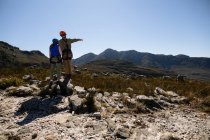 Vue arrière du couple caucasien profitant du temps dans la nature ensemble, portant un équipement de tyrolienne, debout admirant la vue, l'homme pointant sur une journée ensoleillée dans les montagnes — Photo de stock