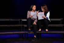 Vorderansicht einer kaukasischen und einer gemischten Rasse Teenager-Mädchen mit Drehbüchern sitzen auf der Bühne eines Schultheaters während der Proben für eine Aufführung — Stockfoto