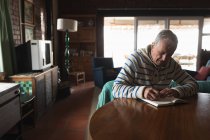 Біля столу сидів старший кавказький чоловік, розслабляючись, сидячи за столом у їдальні і пишучи в книжці з олівцем. — стокове фото